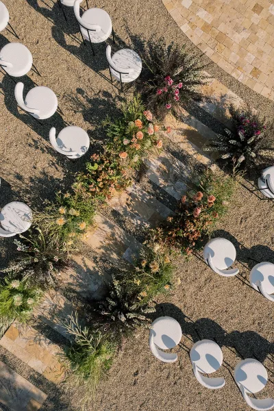 Ceremony aisle decor with aisle floral arrangements lining a stone aisle runner, framed by white chairs on gravel and a stone paver patio