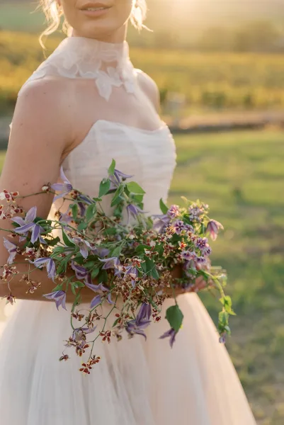 Bridal portrait of a bride holding bouquet of purple wildflowers and greenery, wearing a strapless lace neckline dress in a sunset field