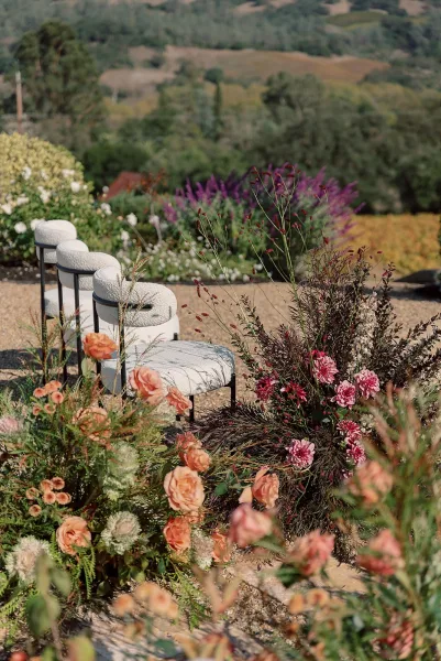 Ceremony aisle decor with outdoor ceremony aisle flowers along a stone path, featuring upholstered stools and peach blooms on a hillside setting