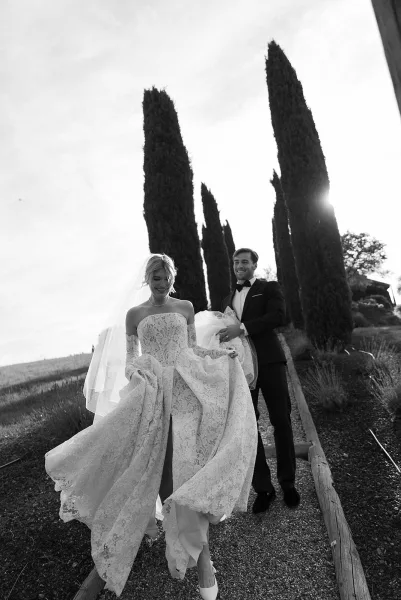 Wedding couple portrait of bride and groom walking as the groom holds her dress train, sunlight on a gravel path lined with cypress trees