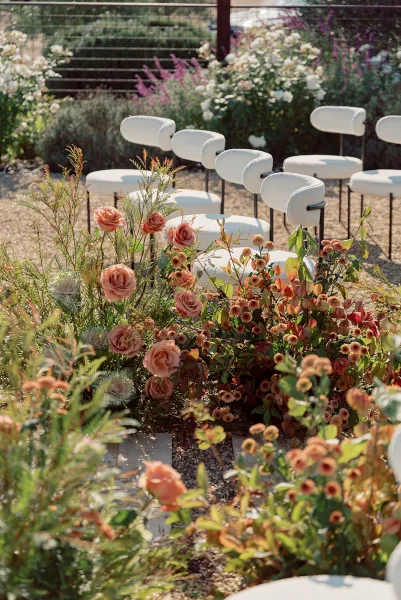 Ceremony setup with outdoor ceremony seating, white chairs lining a gravel aisle with peach rose floral markers and greenery in a garden setting