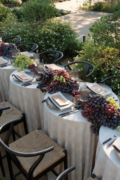 Reception tablescape with an outdoor reception table set in white linen, smoked glass tumblers, and grape centerpiece on a sunlit patio walkway