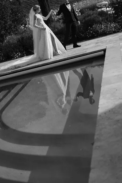 Couple portrait of bride and groom holding hands by a pool, bride in veil and lace gloves, tuxedo groom, hillside garden backdrop