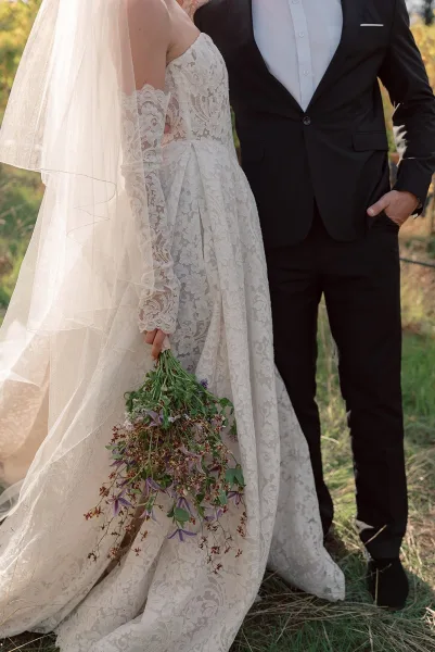 Couple portrait of bride and groom pose, her lace long-sleeve gown and veil with bouquet beside his black suit in golden field light