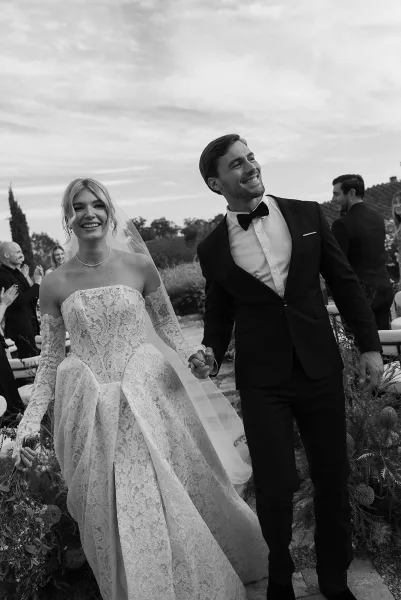 Wedding recessional as bride and groom walk aisle holding hands, bride with bouquet and veil, guests seated along a stone garden path