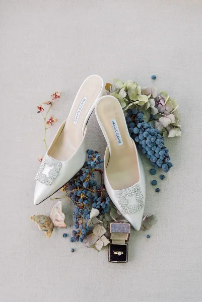 Bridal shoes in a wedding shoe flat lay with white pointed-toe heels and crystal buckles beside a velvet ring box on gray fabric backdrop