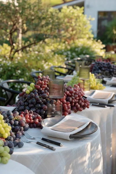 Reception tablescape with outdoor reception table details, grapes centerpiece, striped napkins, ribbed glass votives and candles on linen patio table amid trees