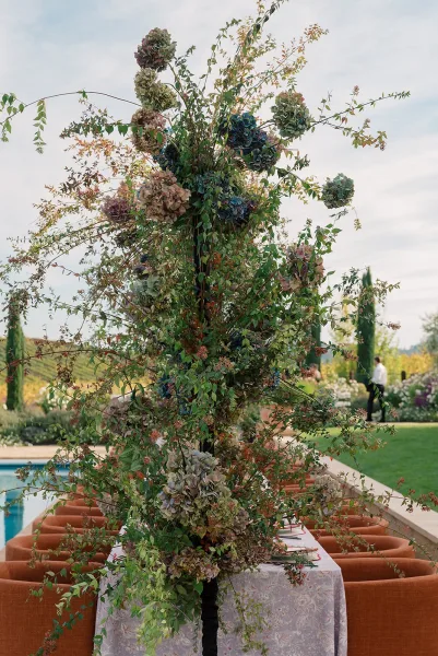 Wedding table centerpiece with a tall floral centerpiece of hydrangeas and greenery branches on a patterned cloth, set poolside on a garden lawn