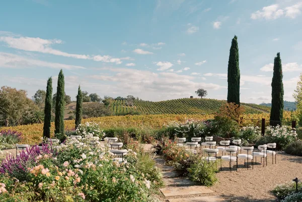 Ceremony setup with white ceremony chairs flanking a stone aisle lined with garden florals, set against vineyard hills and cypress trees