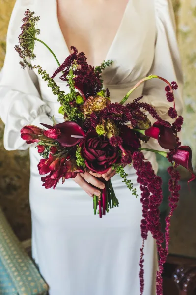 Bridal bouquet in burgundy wedding bouquet tones with calla lilies, ranunculus and cascading amaranthus against a neutral wall