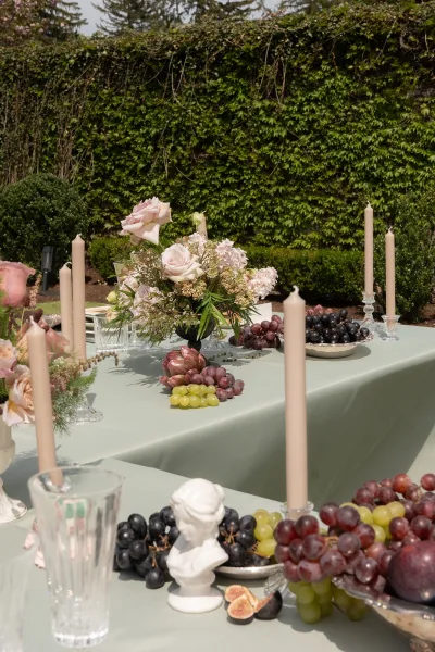 Wedding tablescape at a garden reception table, centered by glowing taper candles, set before a lush ivy wall with trees and trimmed shrubs.