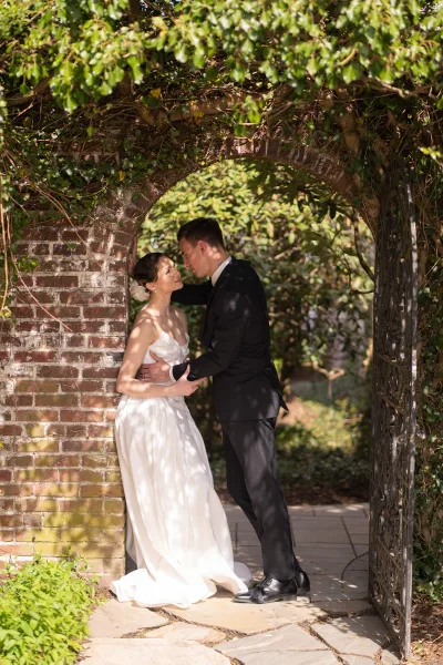 Couple portrait as bride and groom embrace beneath a brick archway and wrought iron gate, with vines, on a stone garden path.