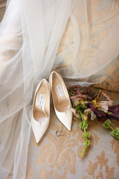 Bridal shoes, ivory wedding heels, rest on a damask upholstered chair with a soft veil draped gently nearby.