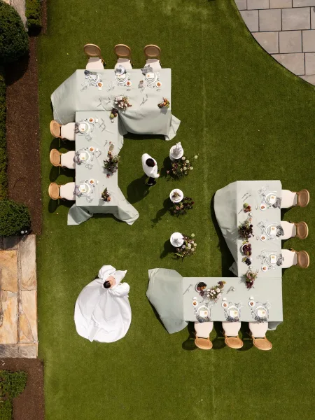 Reception layout with U-shaped wedding tables arranged on a grass lawn, featuring floral centerpieces and framed by hedges and garden beds.