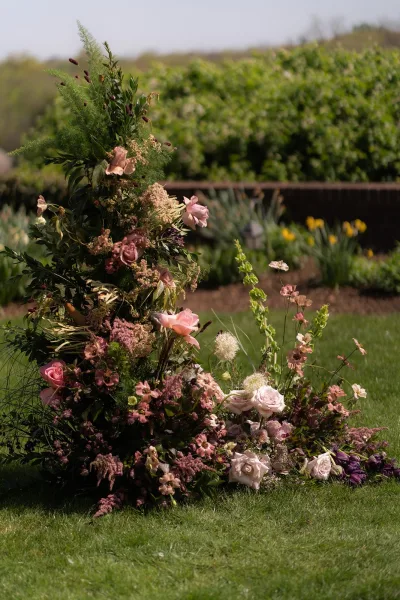 Ceremony floral arrangement styled as a ground floral arrangement with roses on a grass lawn before a brick wall and shaded garden shrubs.