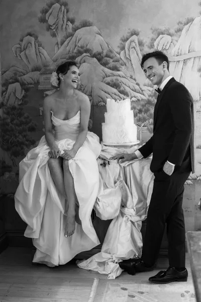 Couple portrait of bride and groom laughing, standing by a wedding cake, framed by a mural wall and wood floor.