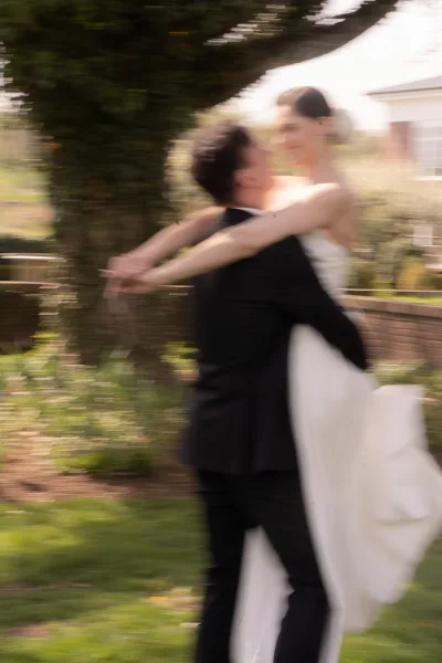 Couple portrait as the groom lifts the bride in a bridal gown on a garden lawn, framed by trees and a brick wall of a building exterior.