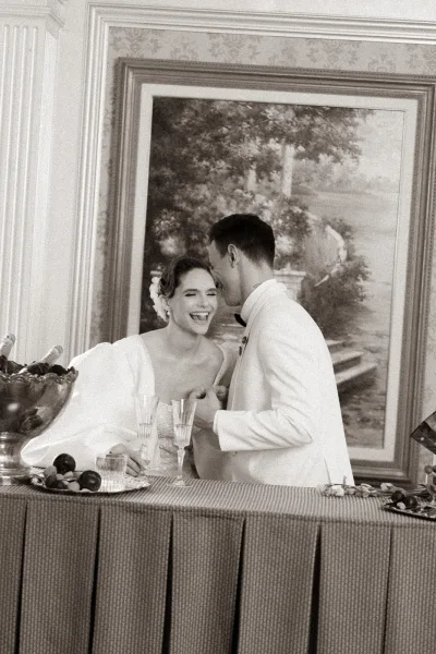 Reception moment with a bride laughing candidly as the groom whispers, toasting with champagne flutes by an indoor bar.