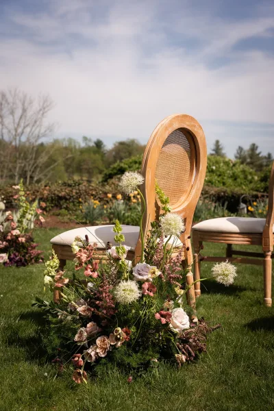 Ceremony aisle flowers arranged as a soft aisle meadow arrangement flanking cane-back chairs, lining the path.