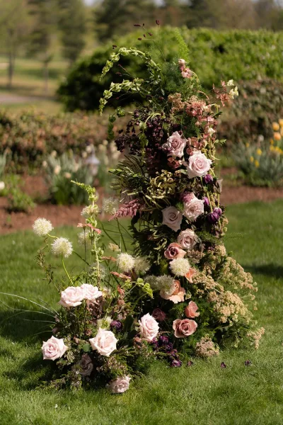 Ceremony floral installation of airy pillars creating a floral pillar wedding backdrop, accented with roses against garden hedges and trees.