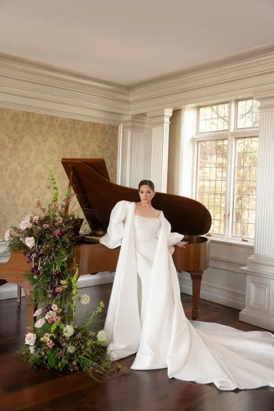Bridal portrait of a bride in a square neck wedding dress, posed beside a grand piano near large windows and white columns on a hardwood floor