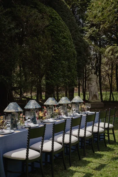 Reception tablescape with blue tablecloth on a long banquet table, floral centerpieces and candles on a garden lawn by a statue