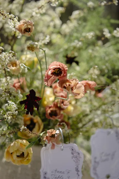 Wedding floral arrangement with spring wedding flowers, featuring tulips and wildflowers beside a place card on a metal ring clip in sunlit garden greenery