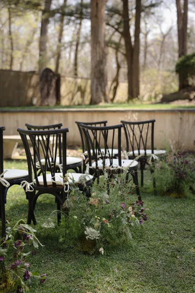 Ceremony aisle decor with black chairs and white cushions, ribbon ties, and ground florals with greenery along a garden lawn by a stone wall