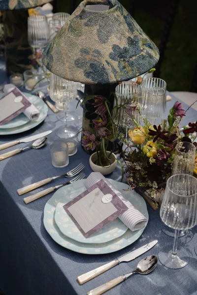 Reception tablescape with wedding table centerpiece of a glowing table lamp, wildflowers, taper candles, and blue linen tablecloth amid garden greenery
