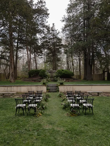Ceremony setup with black chairs and aisle greenery garland leading up stone steps, flanked by benches on a lawn under tall trees