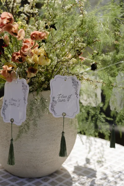 Escort cards hanging from metal hooks on a stone vase, black calligraphy on white cards with peach and yellow flowers in a sunlit garden