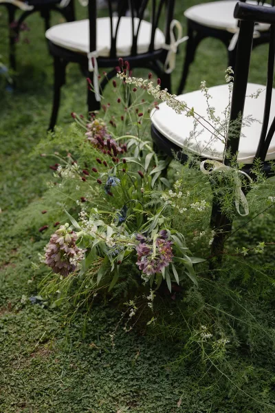 Ceremony aisle florals with aisle ground florals of white and purple wildflowers and greenery along black chairs on a grass lawn outdoors