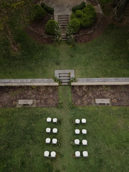 Ceremony setup with white chairs and aisle greenery leading down a brick walkway on a garden lawn with shrubs, stone steps, and benches