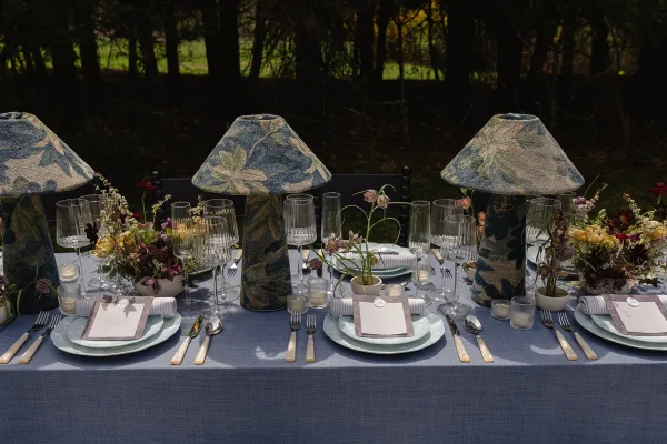 Reception tablescape with outdoor reception table setting, floral lampshade centerpiece, candles, place cards, and glassware on a garden lawn