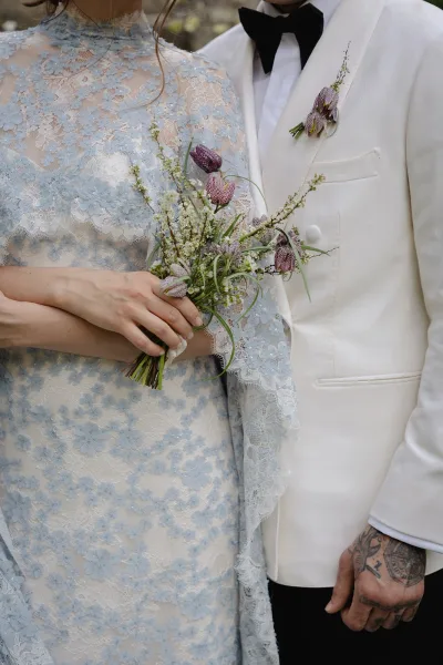 Couple portrait with bride and groom close up, her high-neck long-sleeve lace dress and purple bouquet beside his white tuxedo jacket in soft greenery