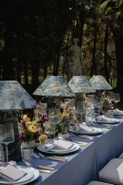 Reception tablescape with outdoor wedding table setting on a blue tablecloth, patterned lamps, wildflower centerpieces, and votive candles in a sunny garden.