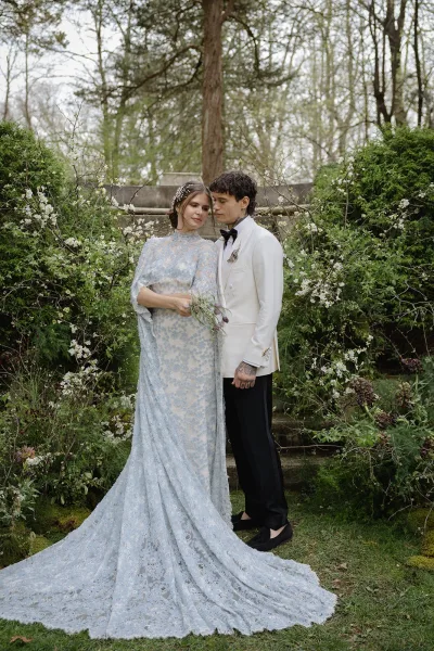 Couple portrait in a garden, bride in a lace long-sleeve gown holding a bouquet beside groom in white tuxedo on stone steps