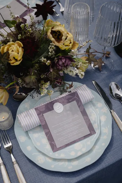 Reception tablescape with wedding place setting featuring patterned plates, striped napkin, wax-sealed menu card, tulip centerpiece on blue linen