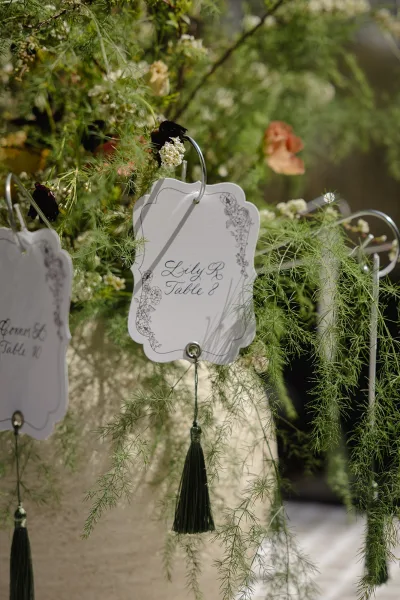 Escort cards on metal hooks with calligraphy and black tassel accents, surrounded by greenery and flowers against a stone wall outdoors