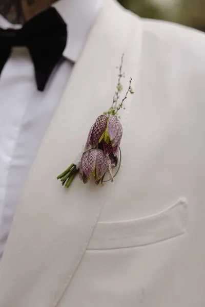 Groom boutonniere with purple boutonniere bell flowers and greenery pinned to a white dinner jacket, black bow tie, blurred garden backdrop