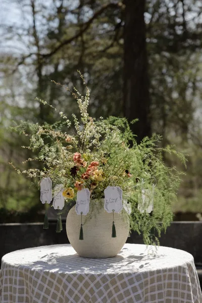 Wedding table centerpiece with a wedding floral centerpiece in a large stone vase, greenery and escort cards on lace tablecloth outdoors in sunlight