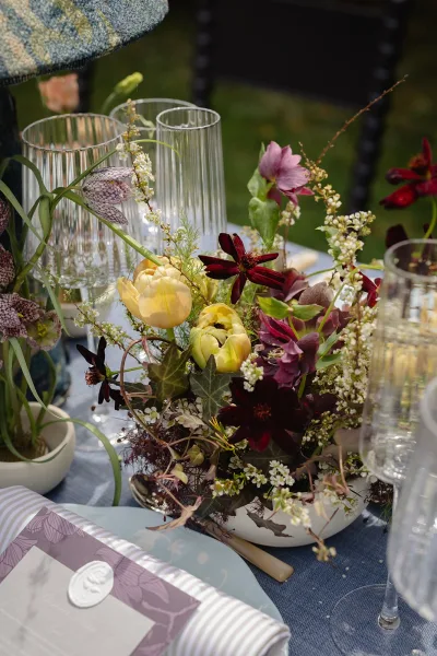 Reception tablescape with wedding floral centerpiece, ribbed glass candle holders, and wax-sealed napkin on blue linen outdoors on a lawn
