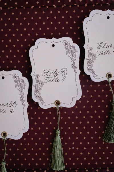 Escort cards with scalloped paper tags and calligraphy, tied with string and tassels, displayed on a patterned fabric backdrop