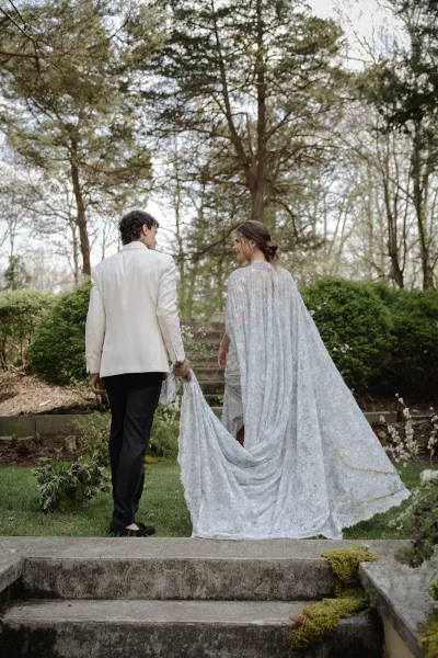 Couple portrait of bride and groom walking away in a garden, groom holding the wedding dress train as they climb stone steps