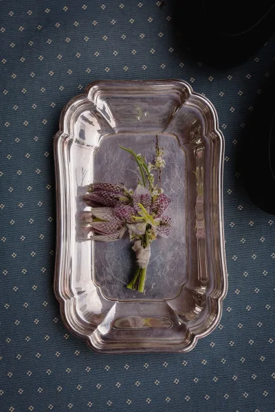Boutonniere detail with small white and purple flowers wrapped in floral tape, styled on a silver serving tray over patterned blue fabric