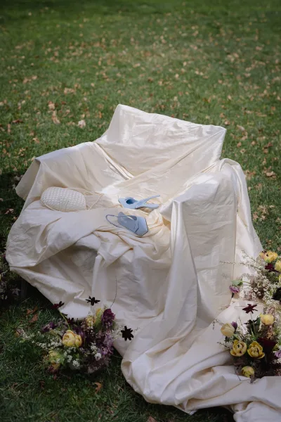 Wedding shoes with something blue heels styled on ivory drape beside woven clutch and florals on grass with scattered leaves