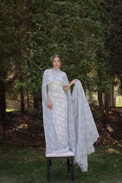 Bridal portrait of a bride in a lace cape wedding dress with high neck and floral appliqué, holding her train on a garden lawn by evergreens