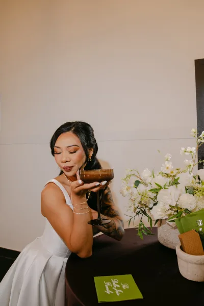 Bridal portrait of a bride sipping cocktail from a coupe glass, with side-swept hair and neutral lipstick highlighting a modern look.