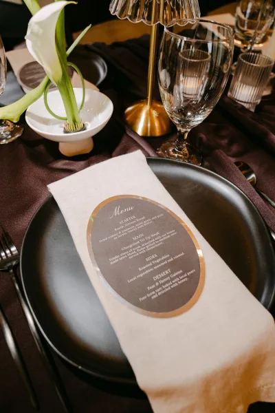 Wedding menu card with oval menu design lies on a satin tablecloth, accented by a single white calla lily in a minimal tabletop setting.