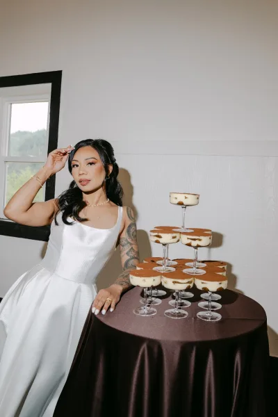 Bridal portrait of a bride with dessert tower, wearing a modern satin wedding dress with a square neckline and soft waves in her hair.